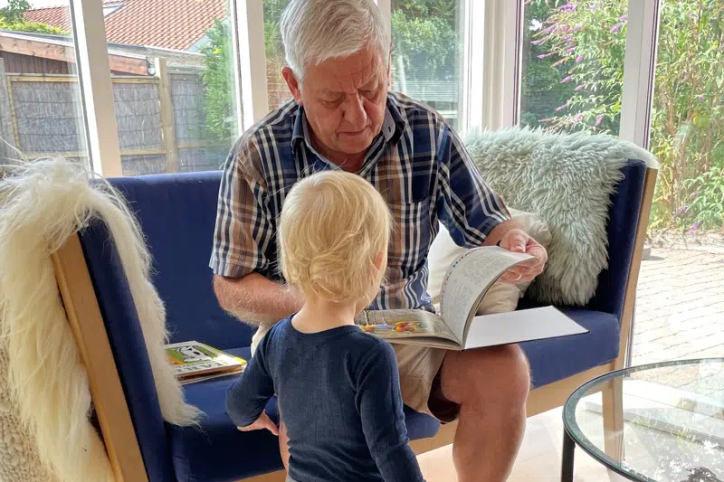 Bedstefar sidder i sofa og læser bog for lille barn.